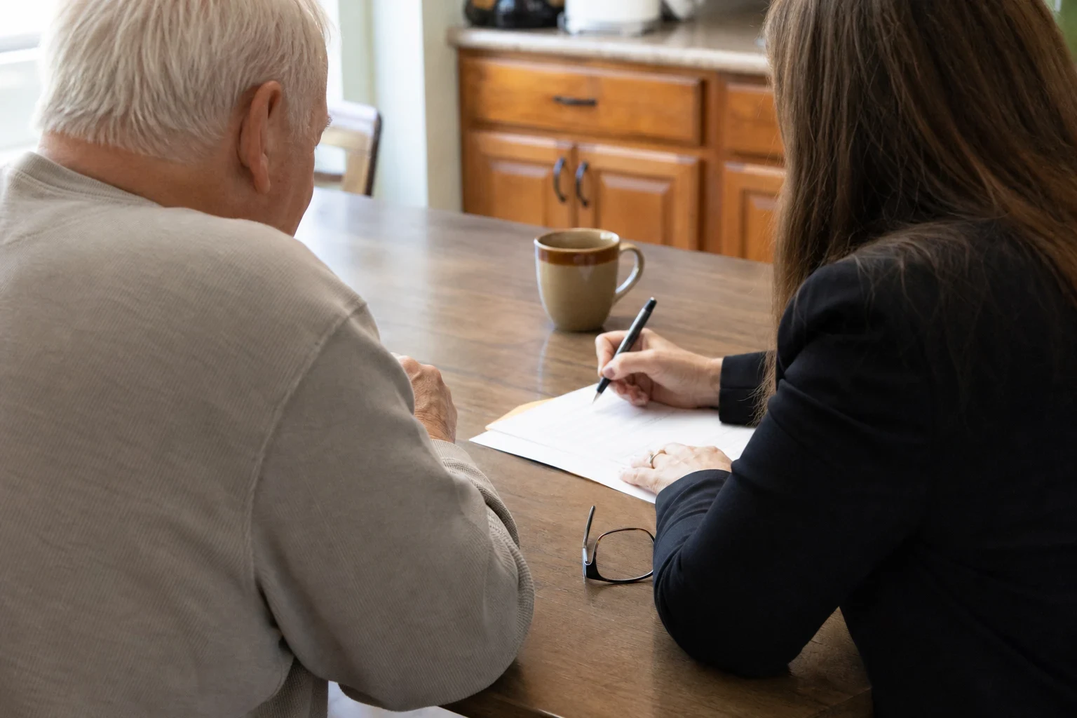 An older man and a woman in a dark blazer sit at a wooden kitchen table reviewing a paper form, with a coffee mug, reading glasses and oak cabinets behind them