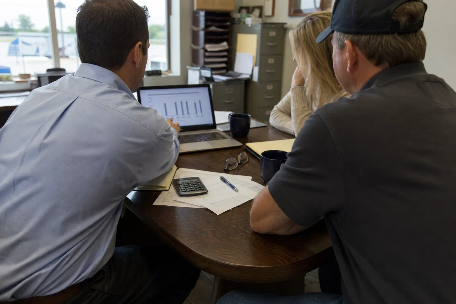 Agent in a light blue button-down shows a bar chart on a laptop to a man in a cap and a woman at a wooden table with papers, calculator and mugs in a small office