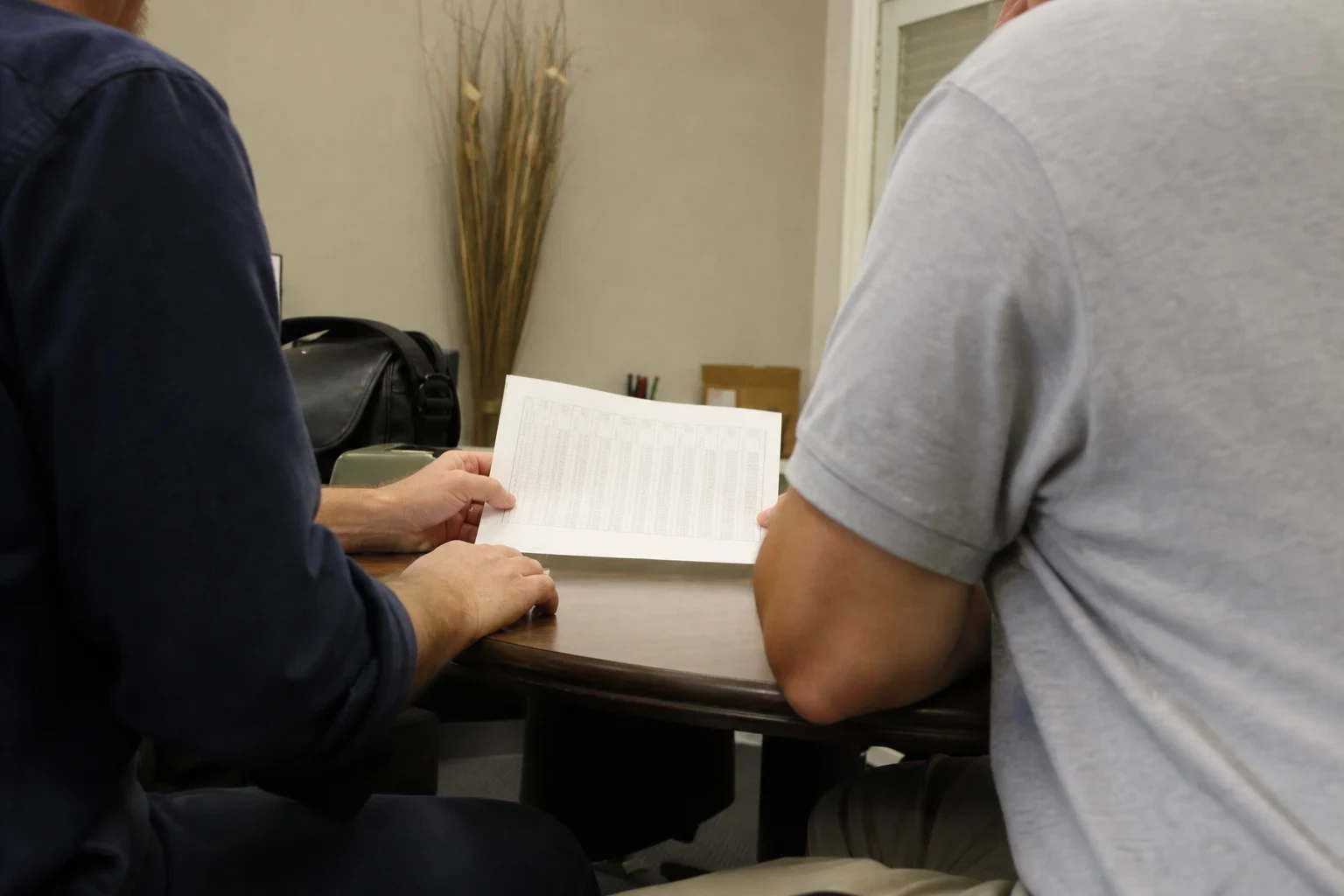Two men seated at a small round wooden table in an office, one holding a printed page of figure tables while both look it over, a black bag and dried grass decor behind.
