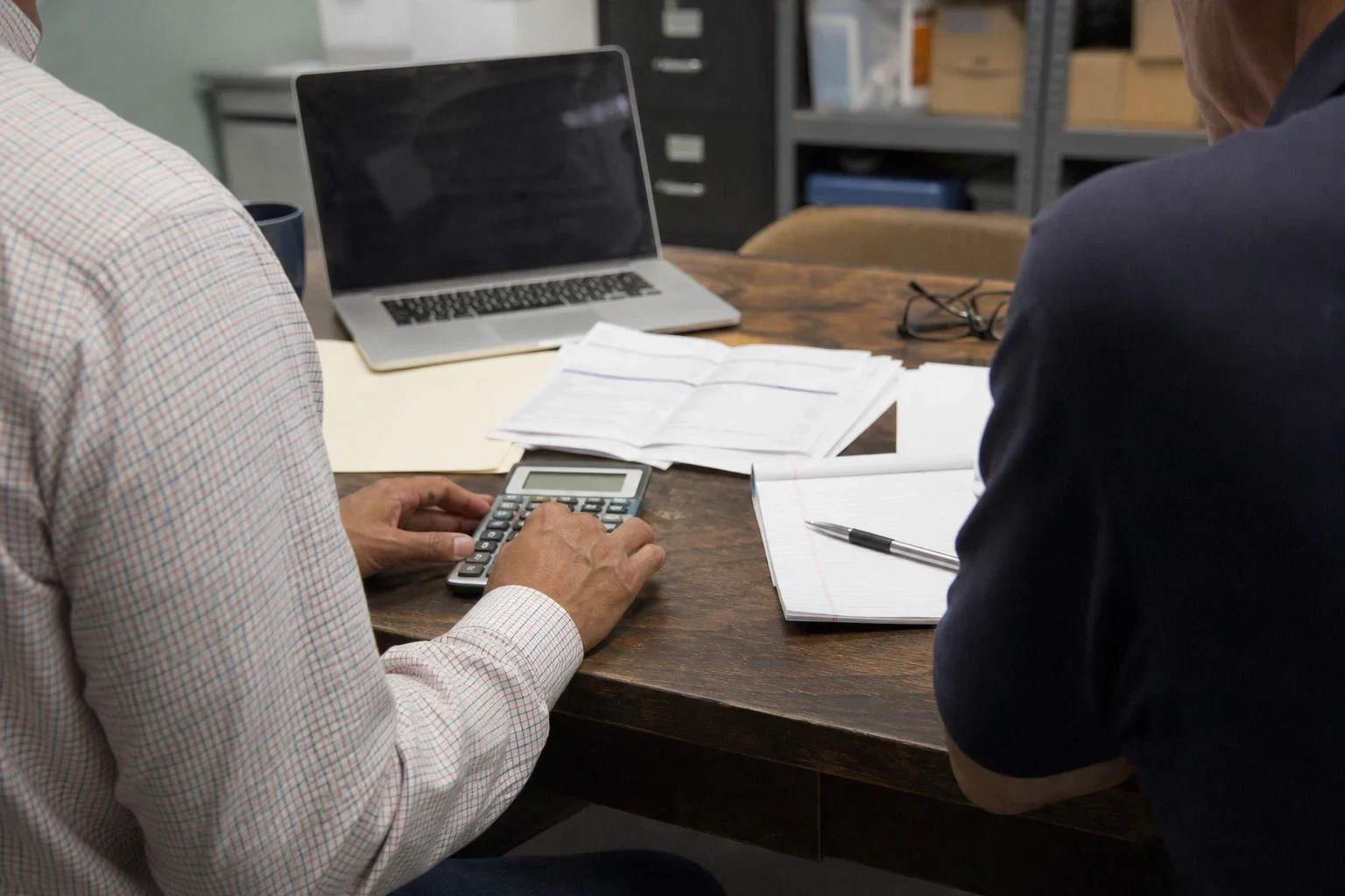 Over-the-shoulder view of two men at a wooden office desk, one using a calculator beside printed statements, a notepad with pen, a laptop and reading glasses.
