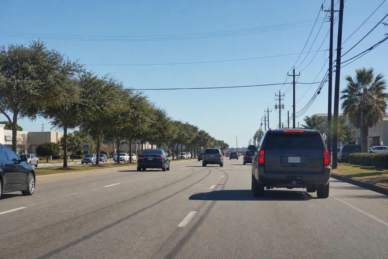Multi-lane suburban road with SUVs and sedans in traffic, a row of live oaks along the median, palm trees, utility poles and low retail buildings under a clear sky